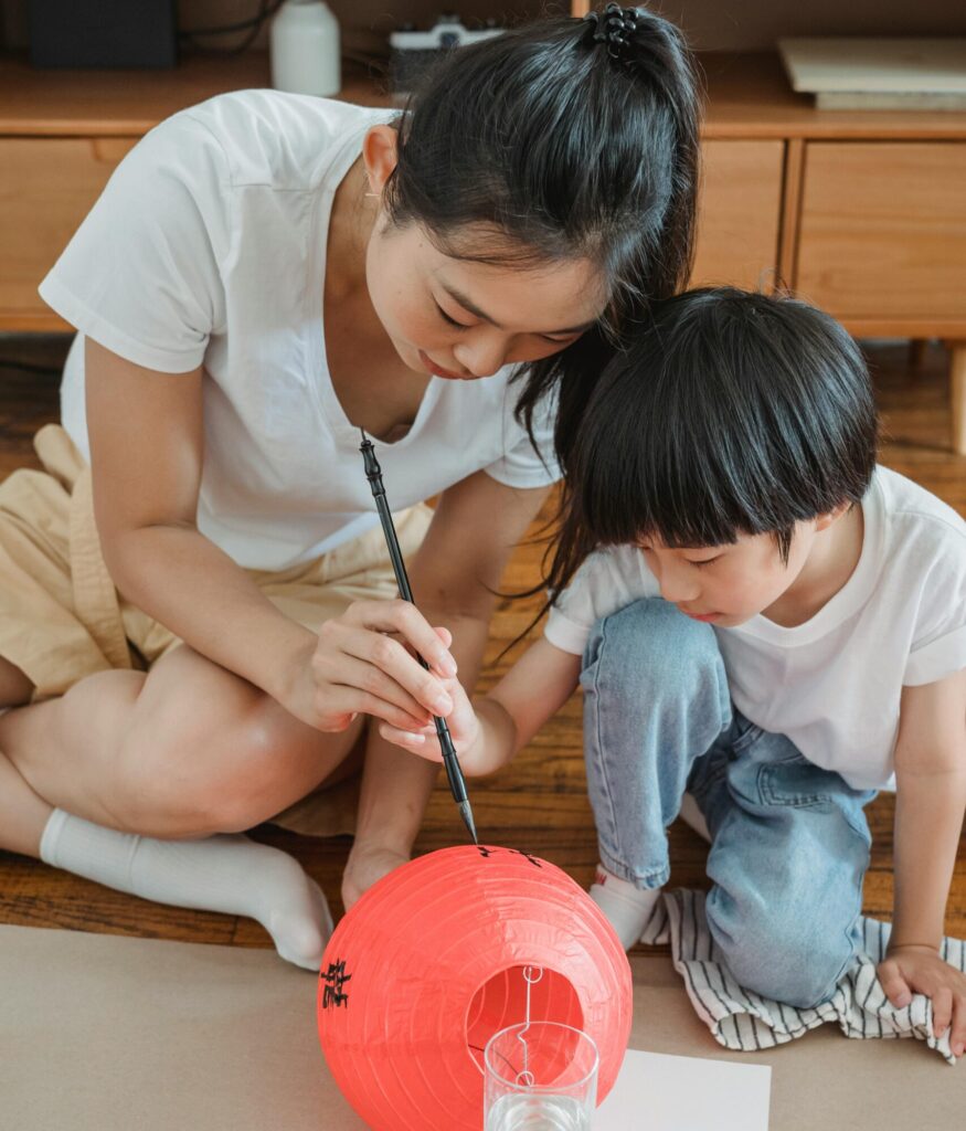 An Asian mother guides her son in painting a bright red lantern indoors, embracing cultural traditions.